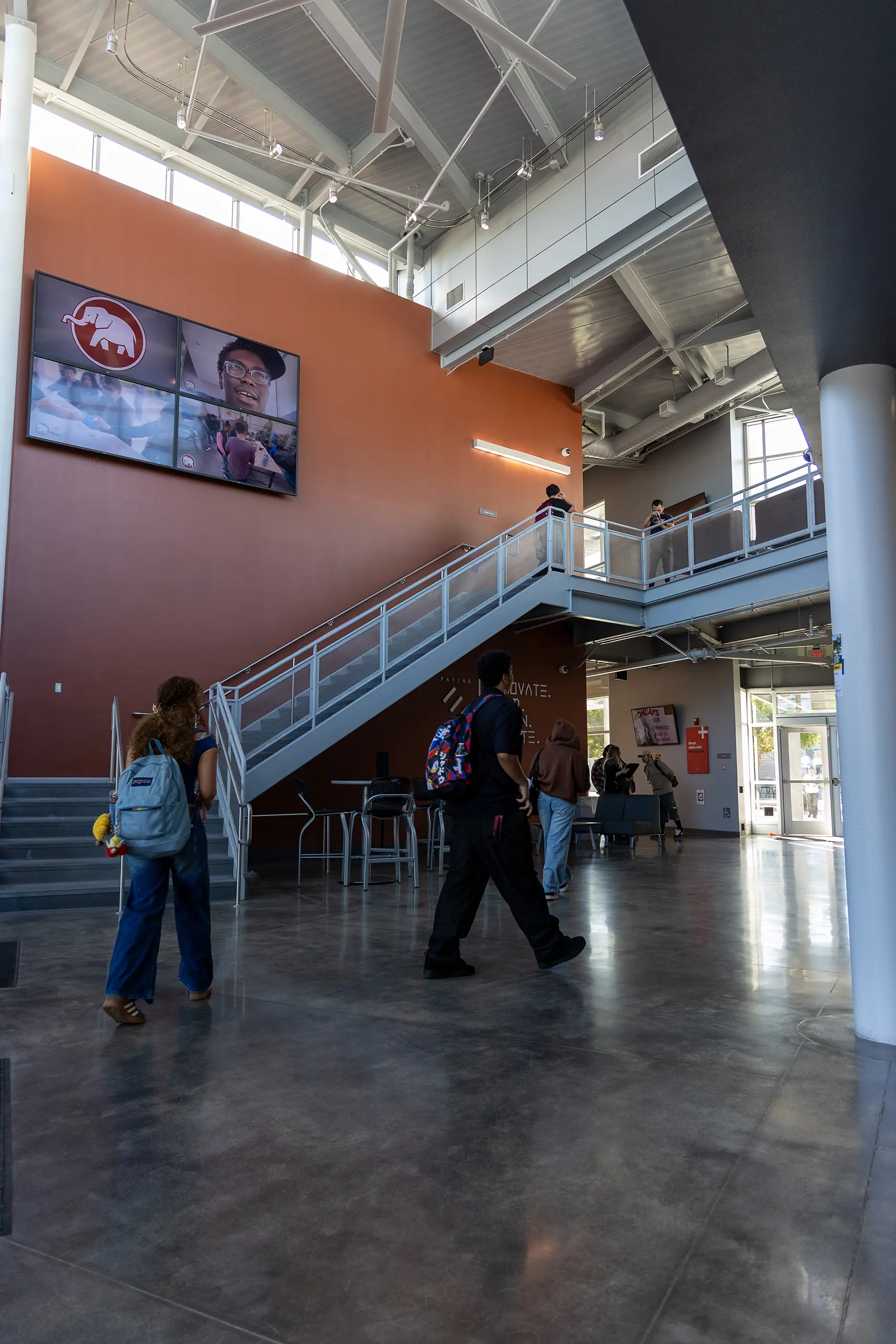 Students walk through a modern building with glass doors, a staircase, high ceilings, and large digital screens on an orange wall showing a logo and a persons face.
