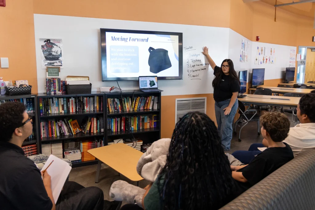 A woman stands by a screen giving a presentation to four seated people in a room with bookshelves, taking notes and listening. The screen displays Moving Forward and an image of a black object.