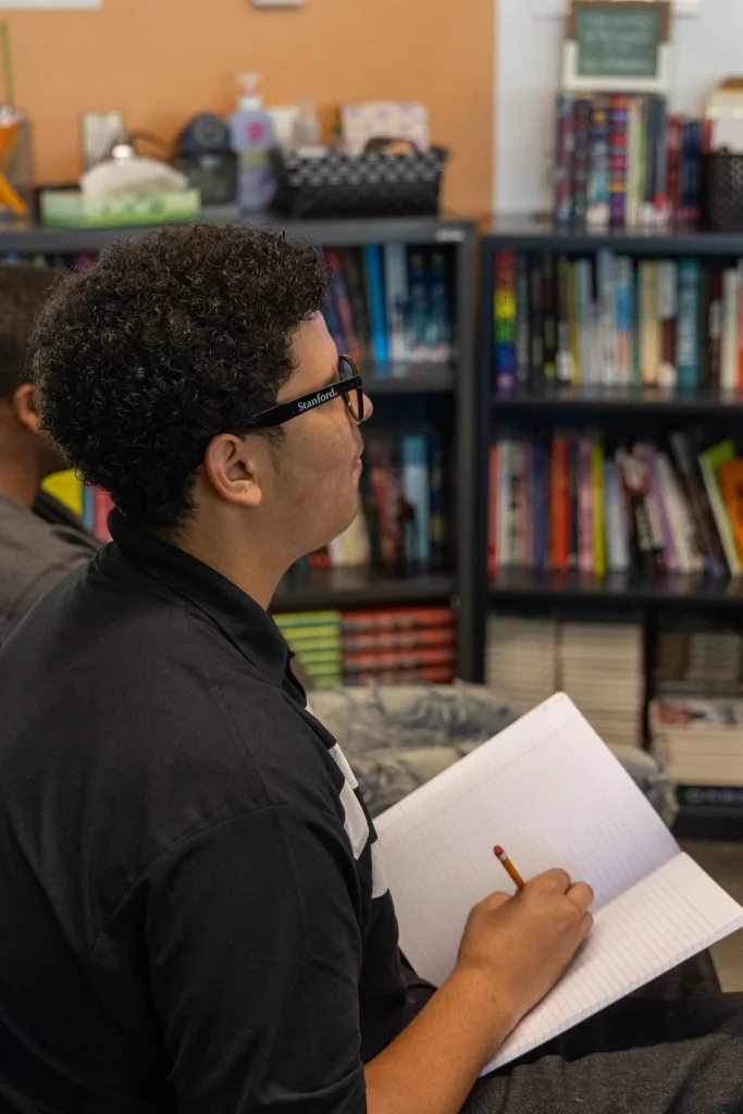 A student with curly hair and glasses sits in a classroom, holding a pencil and an open notebook, appearing to take notes. Bookshelves filled with books are visible in the background.