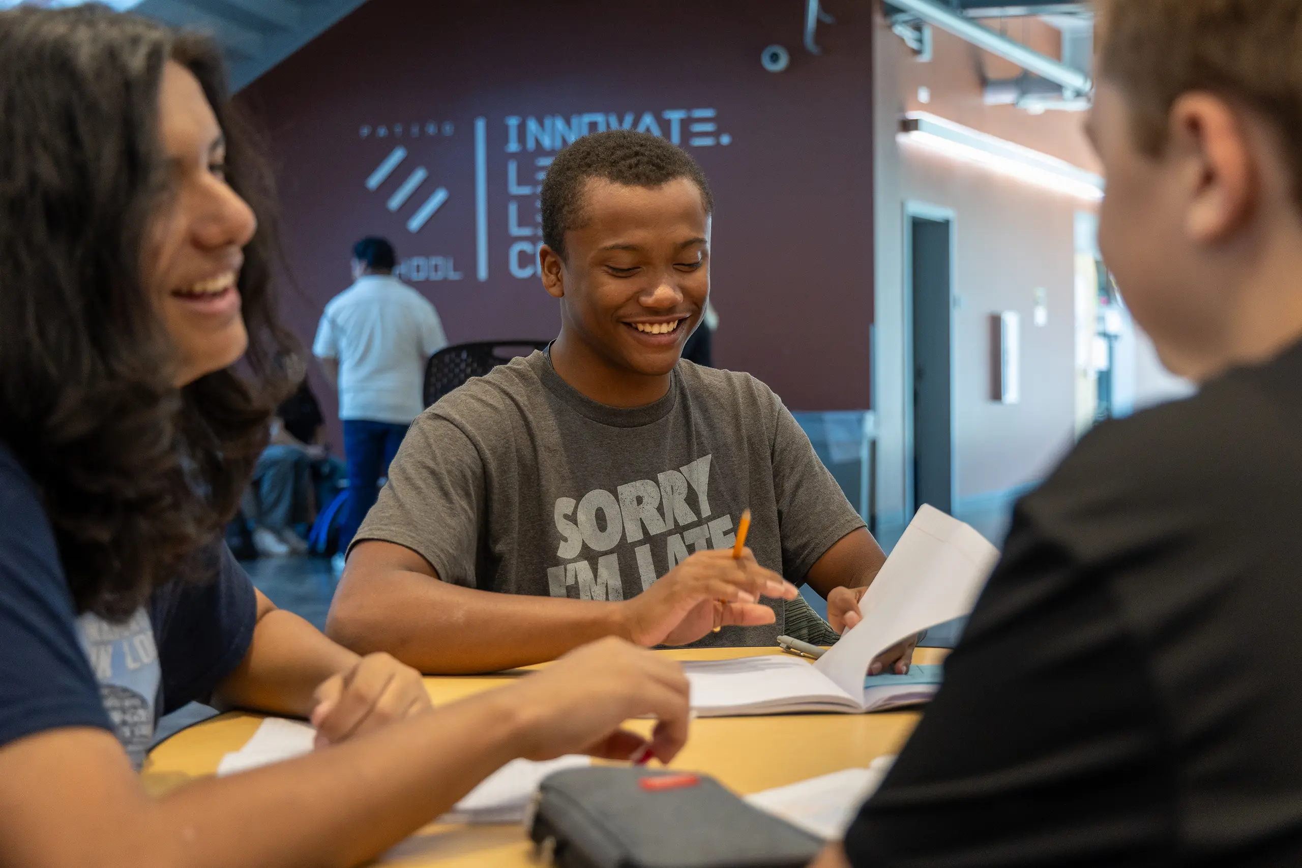 Three students sit at a table, smiling and talking while working on assignments. One student holds a pencil and a notebook. They appear to be studying together in a brightly lit, modern space with inspirational words on the wall.
