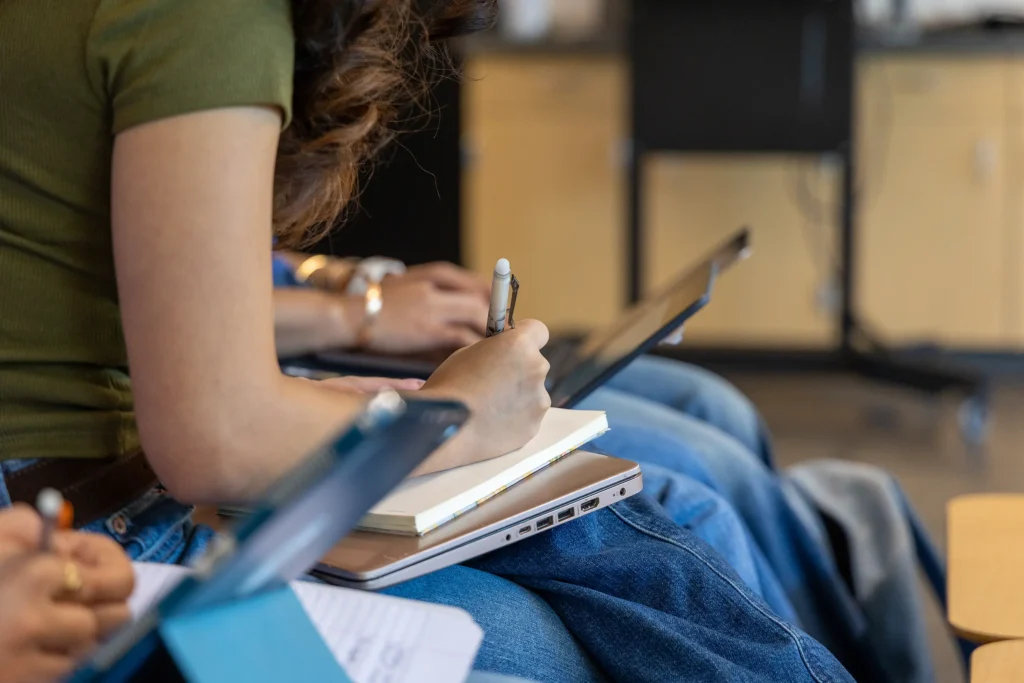 Close-up of students sitting in a row, taking notes on notepads and laptops during a class or lecture. Only their hands, arms, and legs are visible. The focus is on one person writing in a notebook.