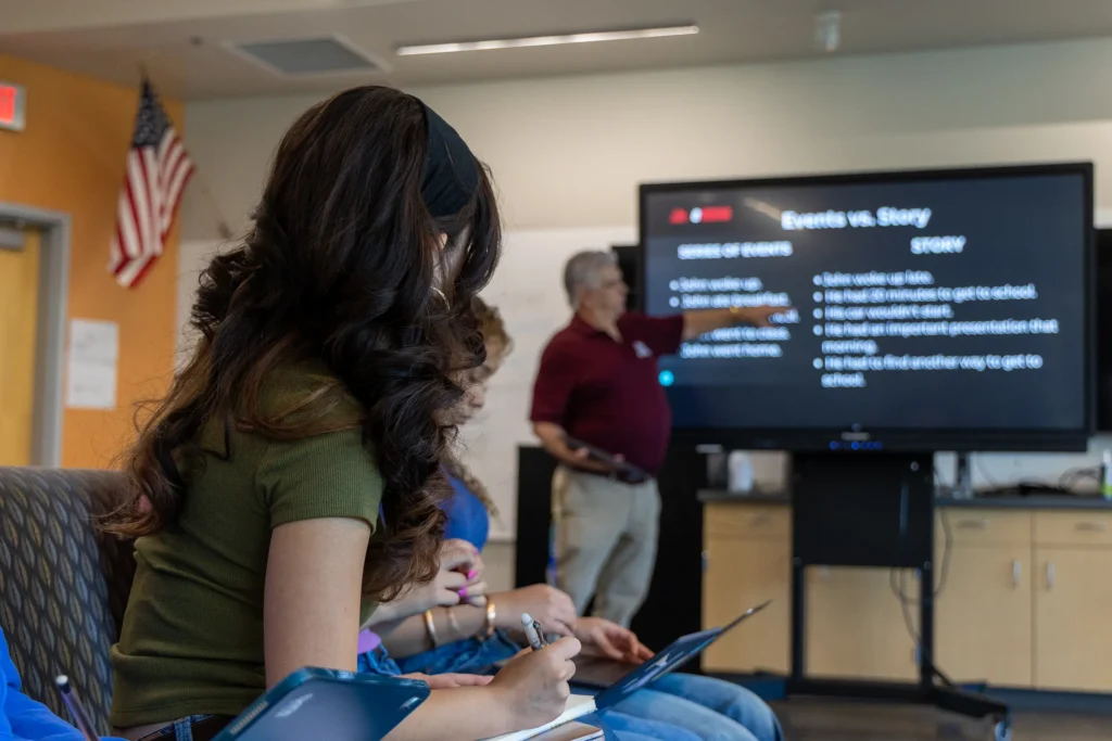 A student takes notes while a teacher stands by a screen displaying Events vs. Story in a classroom. Other students are seated nearby, and an American flag is visible in the background.