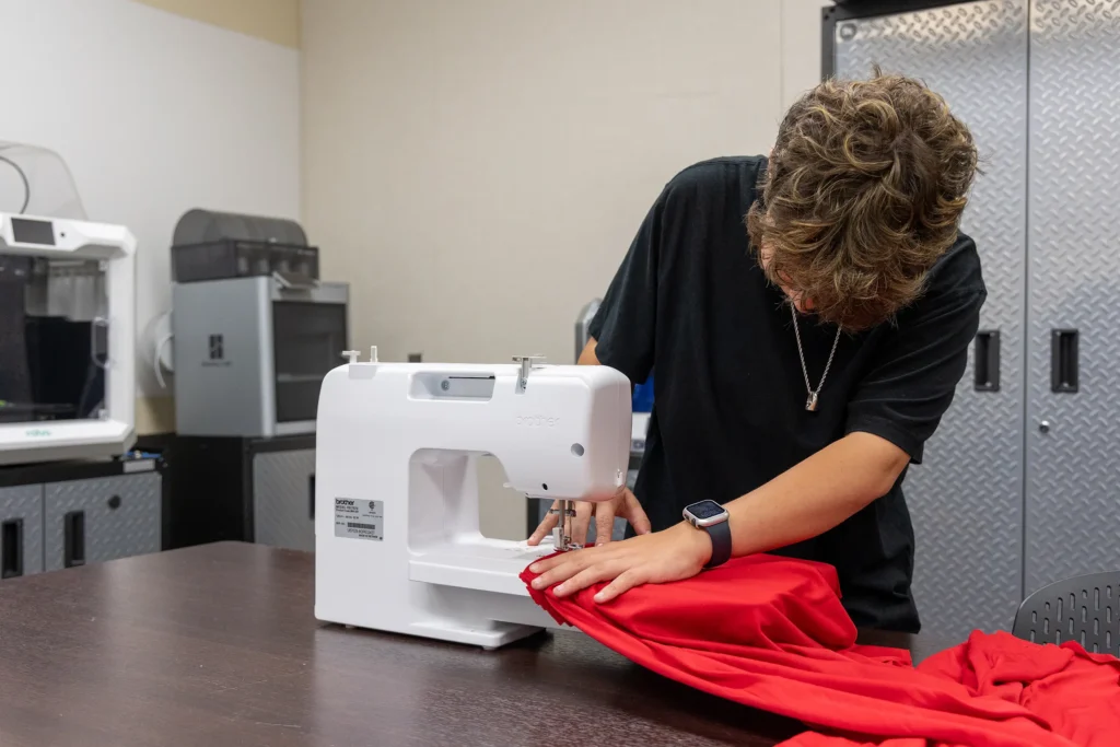 A person with short hair uses a white sewing machine to sew red fabric on a dark table in a room with metal cabinets and large equipment in the background.