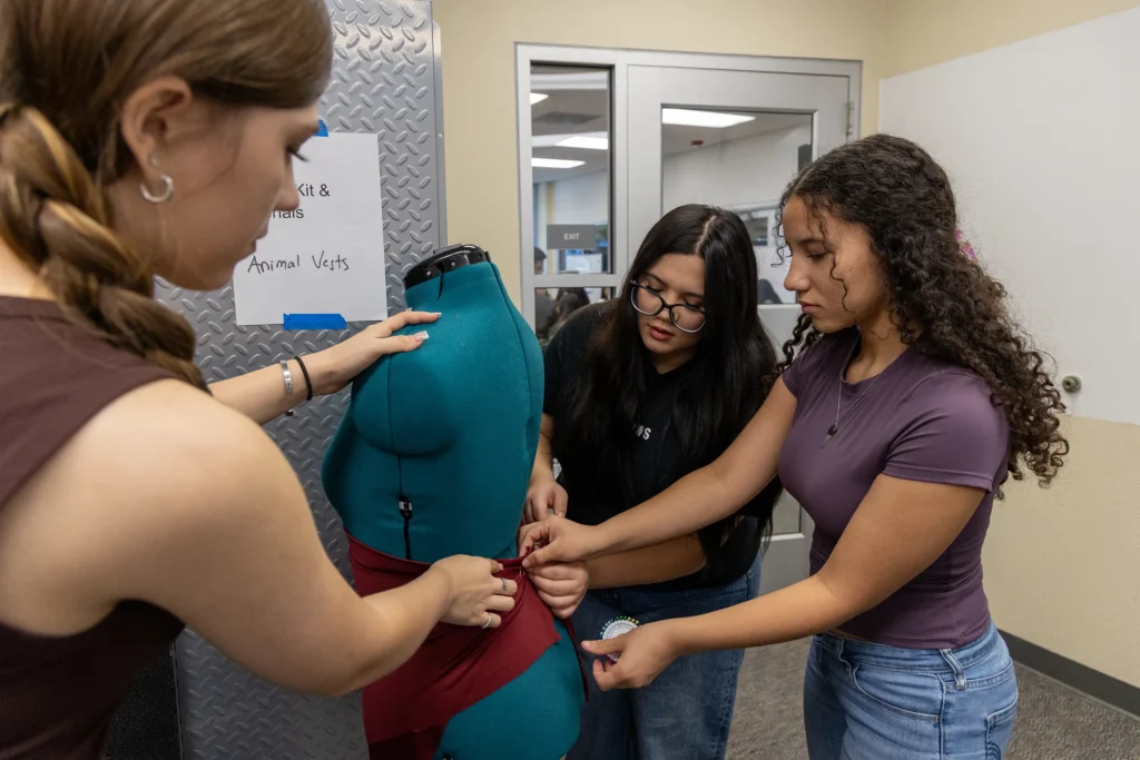 Three young women work together to wrap a red bandage around a teal medical mannequins abdomen in a classroom or lab setting.
