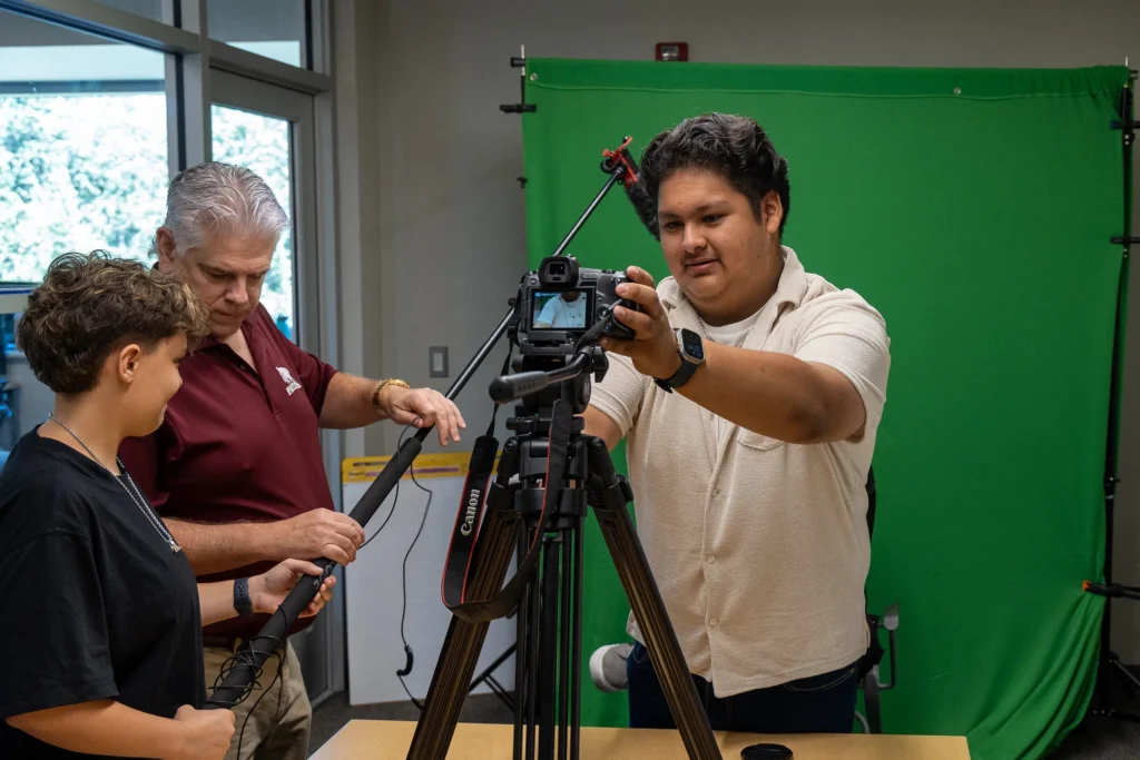 Three people set up a camera on a tripod in front of a green screen in a room, adjusting equipment and cables with a window and whiteboard in the background.