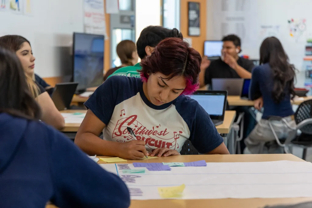 A student with short red hair writes on paper at a classroom desk. Other students in the background use laptops and talk, creating a busy, collaborative learning environment.