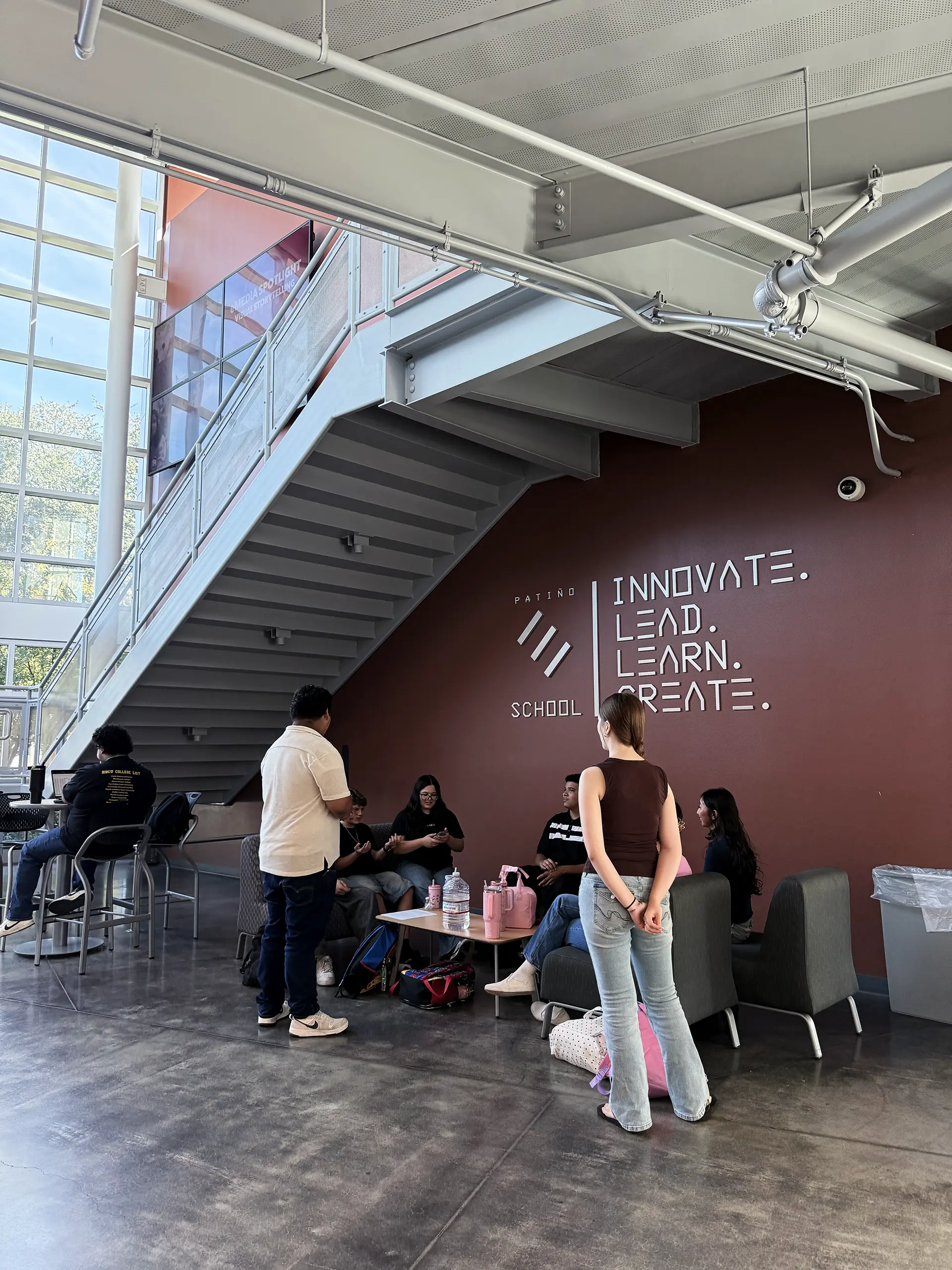 Students gather and talk in a modern school lounge area under a staircase, with Innovate. Lead. Learn. Create. and the schools logo on a red wall. Natural light enters through large windows.