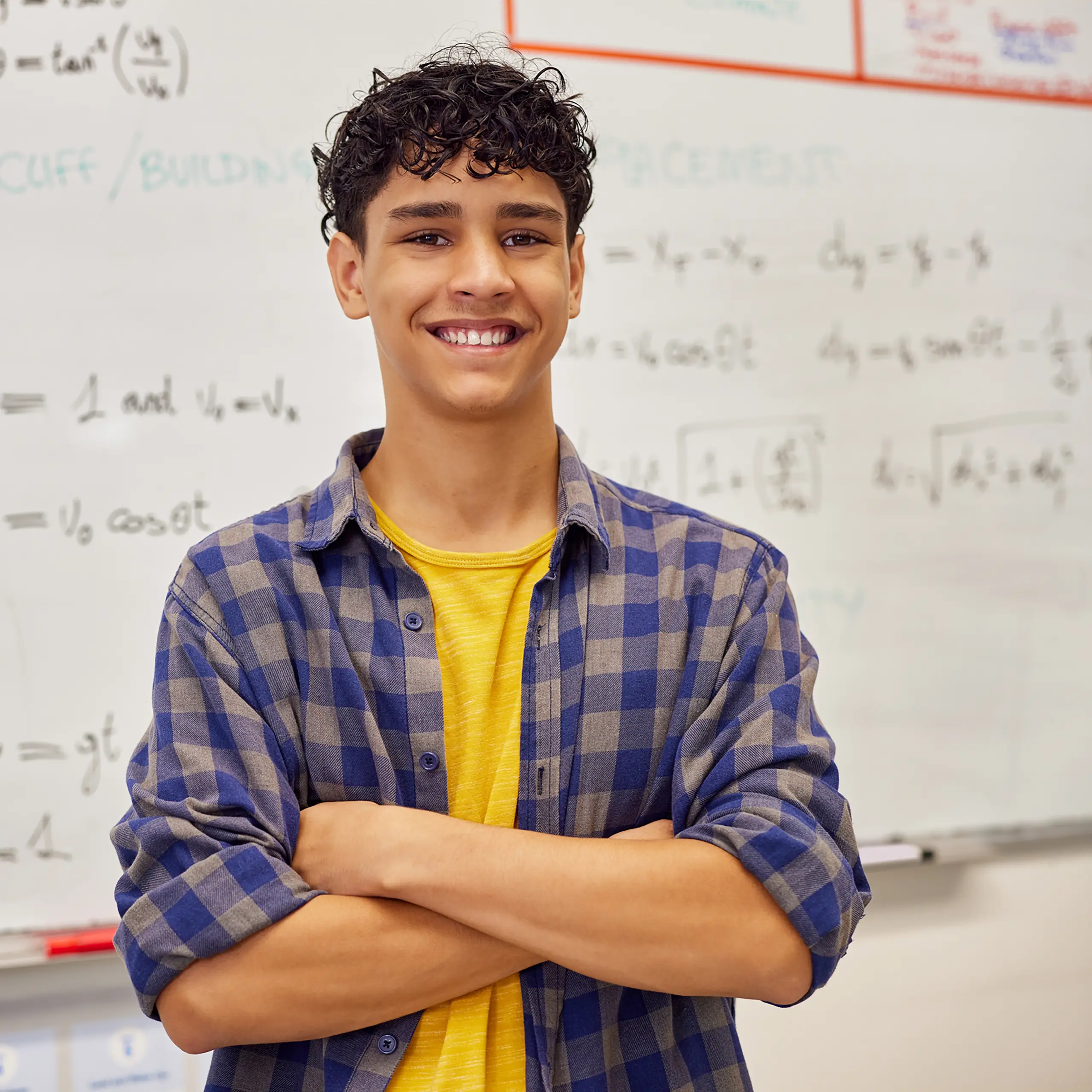 Smiling teenage student with curly hair stands with arms crossed in front of a whiteboard covered in math equations, wearing a yellow shirt and blue plaid button-up.