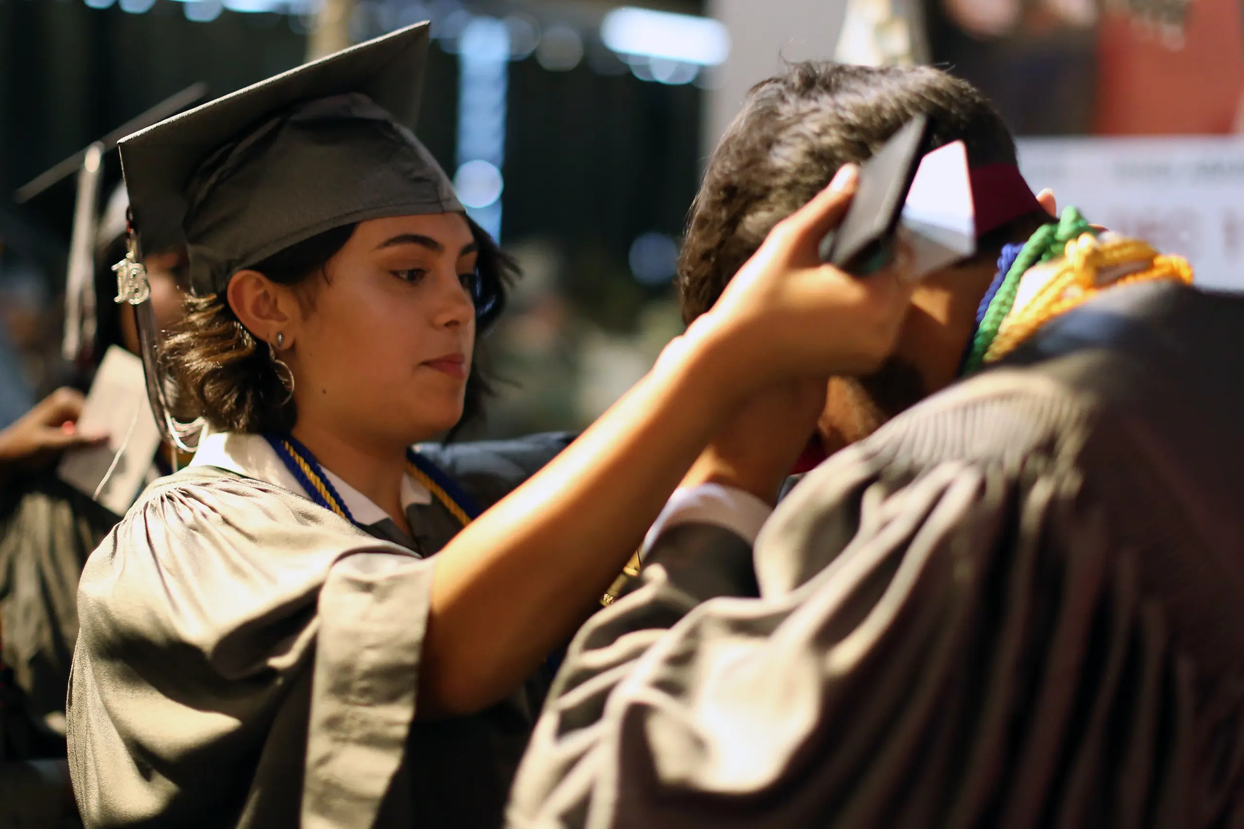 A graduate in a cap and gown adjusts another graduates tassel during a ceremony. The scene is lively, with a blurred background and focus on the act of helping.