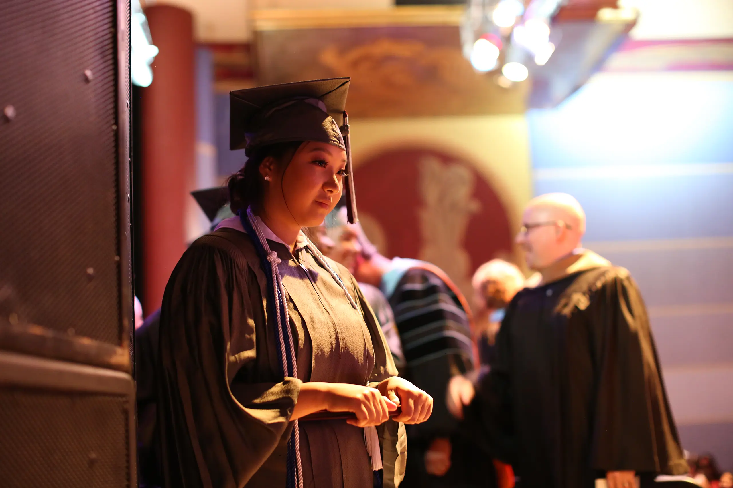 A graduate in cap and gown stands on stage, looking thoughtful, with other graduates and faculty in the background under warm lights.