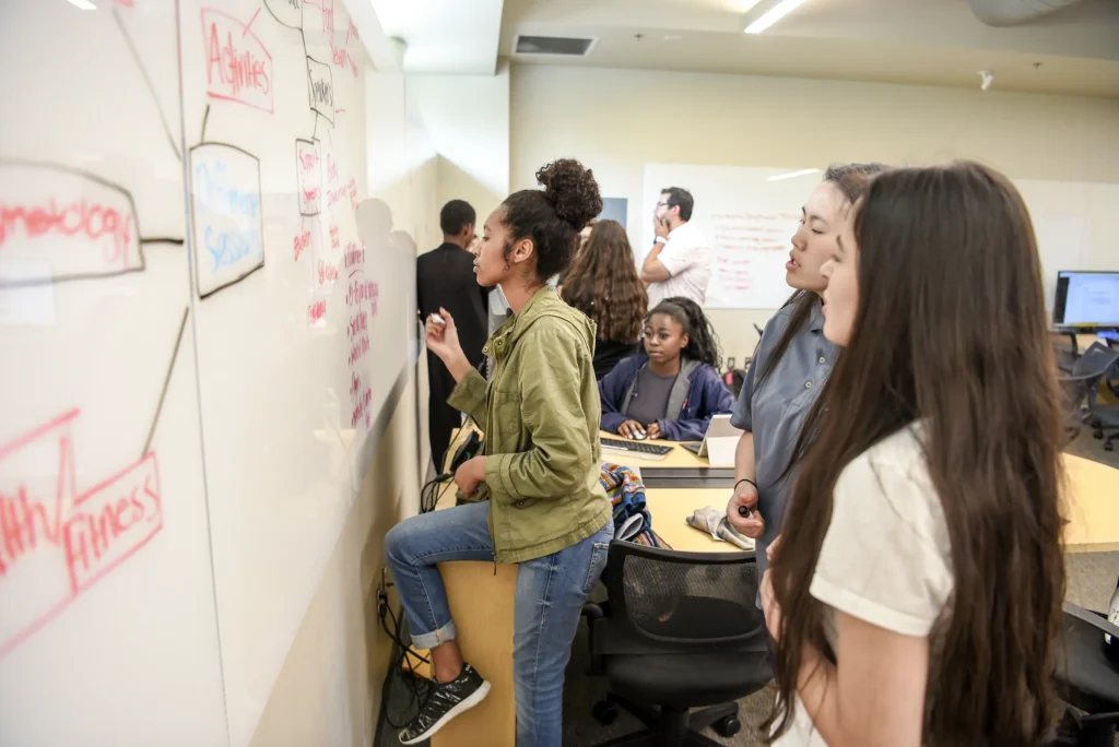 Several students stand at a whiteboard, writing and discussing ideas in a classroom. Other students sit at desks, observing or participating in the group activity.